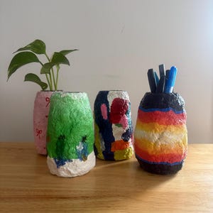 May include: Four handmade, textured vases in a row. One pink vase with a plant, a green vase, a vase with a colorful abstract design, and a rainbow-striped vase holding pens. The vases are on a wooden surface.