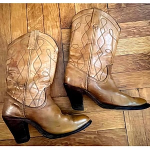 May include: A pair of tan leather cowboy boots featuring a pointed toe, a stacked dark brown heel, and decorative stitching patterns on the shaft. The boots have pull tabs at the top and a smooth finish, showcasing a classic western-inspired design.
