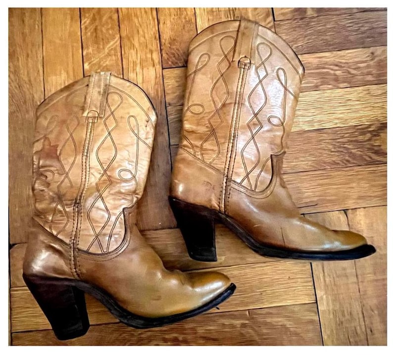 May include: A pair of tan leather cowboy boots featuring a pointed toe, a stacked dark brown heel, and decorative stitching patterns on the shaft. The boots have pull tabs at the top and a smooth finish, showcasing a classic western-inspired design.