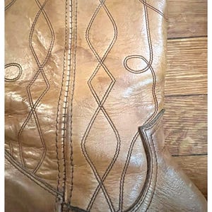 May include: A tan leather boot featuring a vertical seam and decorative stitching in a diamond pattern. The leather has a natural grain texture with visible creases. The brand name FRYE is embossed in small letters near the bottom of the shaft.