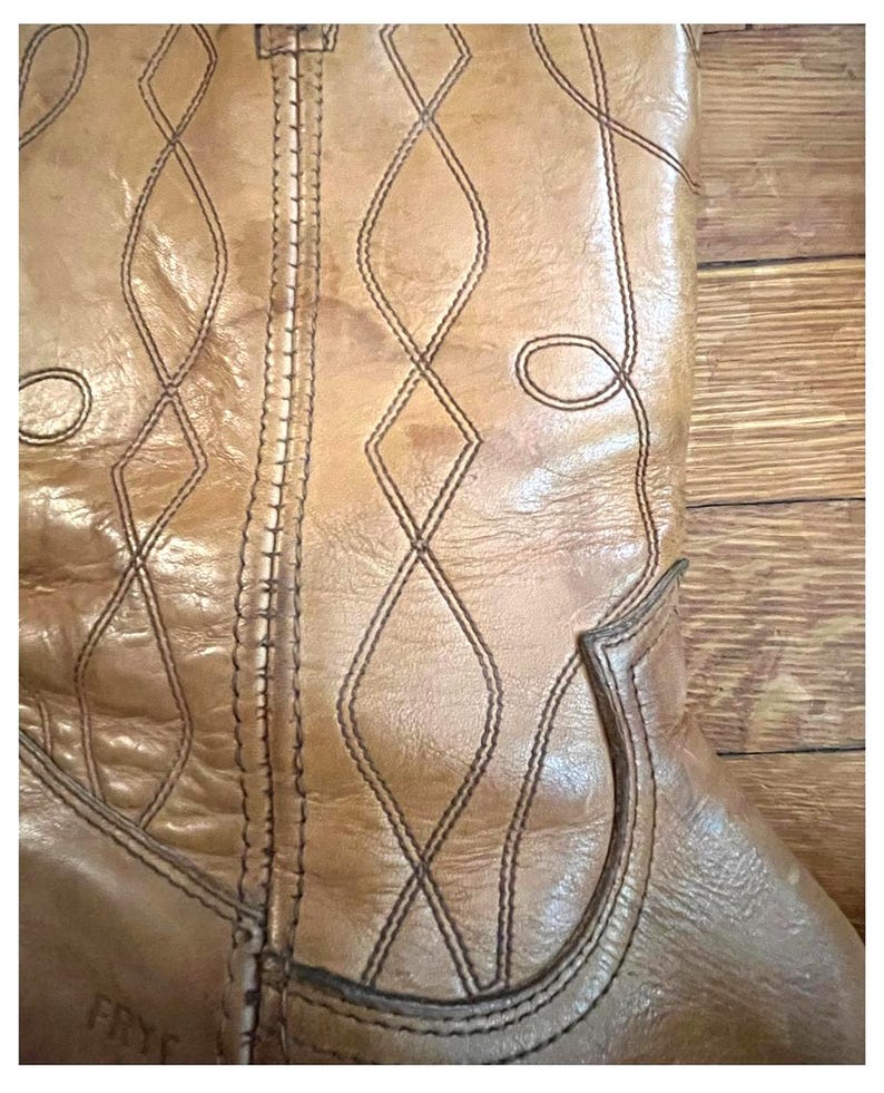 May include: A tan leather boot featuring a vertical seam and decorative stitching in a diamond pattern. The leather has a natural grain texture with visible creases. The brand name FRYE is embossed in small letters near the bottom of the shaft.
