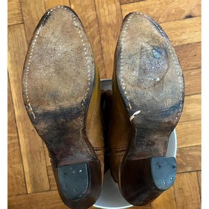 May include: A pair of tan leather boots shown from the bottom view, featuring worn light brown leather soles with visible white stitching along the edges, dark brown stacked heels with black rubber heel caps, and a pointed toe silhouette.