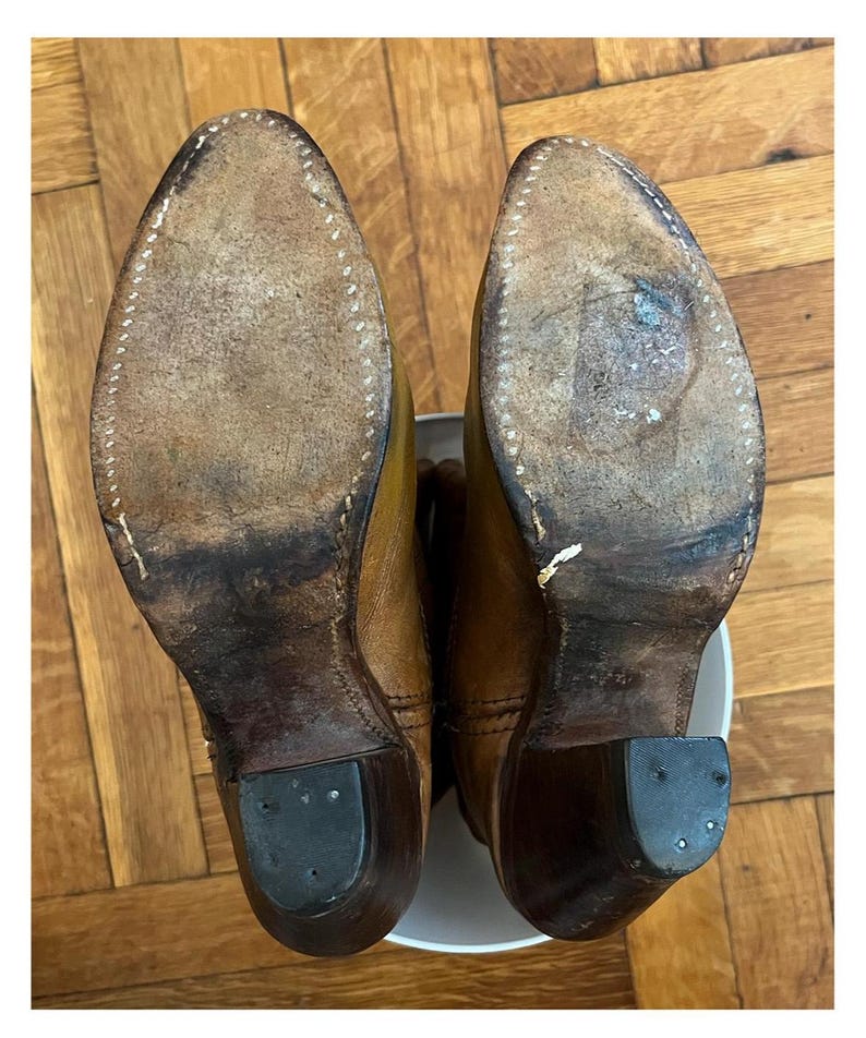 May include: A pair of tan leather boots shown from the bottom view, featuring worn light brown leather soles with visible white stitching along the edges, dark brown stacked heels with black rubber heel caps, and a pointed toe silhouette.