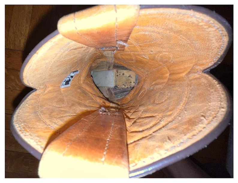 May include: A top-down view into the interior of a brown leather cowboy boot. The lining is a light tan color with visible stitching patterns along the shaft. Two leather pull straps extend upward from the opening, and a small white tag is visible.