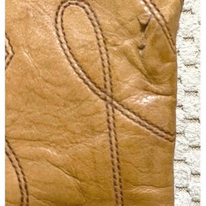 May include: A tan leather boot featuring a textured surface with visible grain and decorative stitching patterns. The stitching forms curved, loop-like designs across the leather material, highlighting the construction and detail of the footwear.