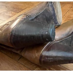 May include: A pair of brown leather cowboy boots featuring a pointed toe, a mid-calf shaft with decorative stitching, and a stacked dark brown block heel. The boots are positioned at an angle, showcasing the leather texture and the heel construction.