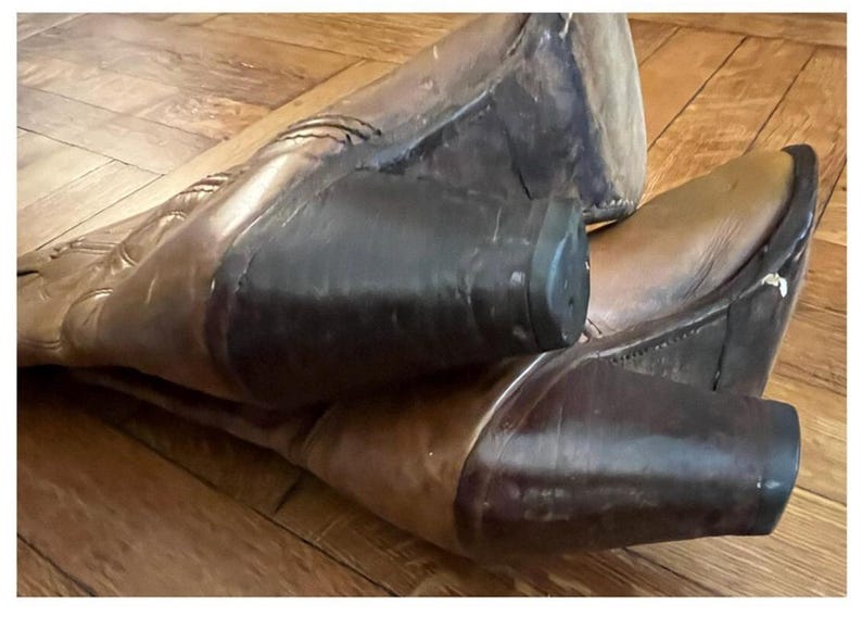 May include: A pair of brown leather cowboy boots featuring a pointed toe, a mid-calf shaft with decorative stitching, and a stacked dark brown block heel. The boots are positioned at an angle, showcasing the leather texture and the heel construction.