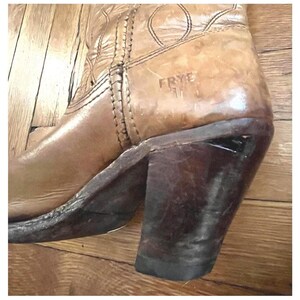 May include: A tan leather boot featuring a stacked dark brown heel and a rounded toe. The side of the boot displays the embossed text FRYE. The upper portion of the boot includes decorative stitching patterns along the seams and leather surface.