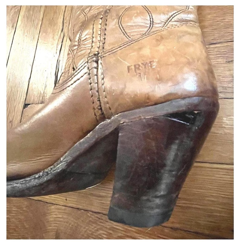May include: A tan leather boot featuring a stacked dark brown heel and a rounded toe. The side of the boot displays the embossed text FRYE. The upper portion of the boot includes decorative stitching patterns along the seams and leather surface.