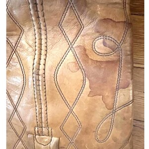 May include: A tan leather boot shaft featuring a vertical seam and decorative stitching in a diamond pattern. The leather surface displays natural variations in color and texture, including a prominent darker brown stain on the right side.