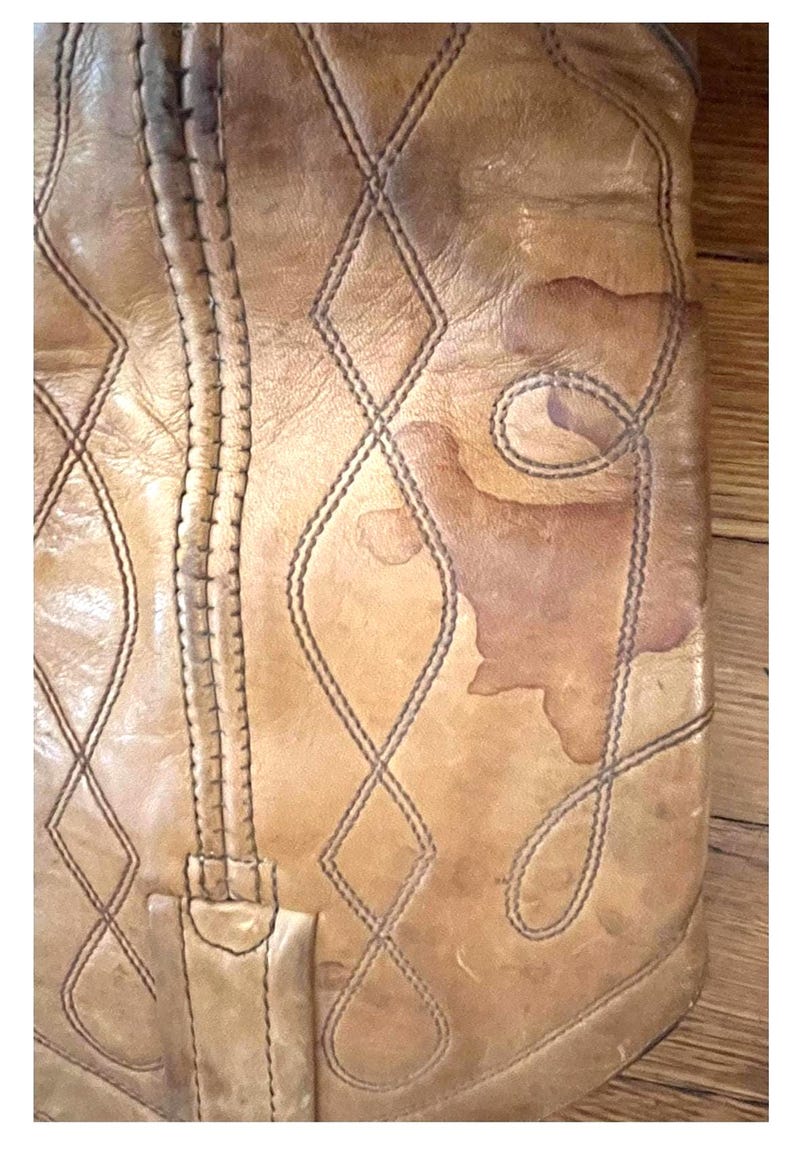 May include: A tan leather boot shaft featuring a vertical seam and decorative stitching in a diamond pattern. The leather surface displays natural variations in color and texture, including a prominent darker brown stain on the right side.