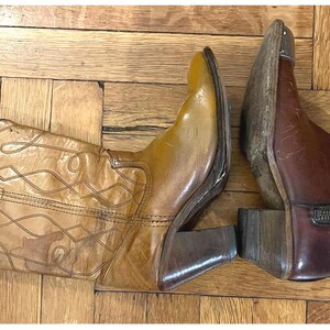 May include: A pair of leather boots consisting of one tall tan western-style boot with decorative stitching and a stacked heel, and one shorter dark brown ankle boot featuring a side zipper closure, a pointed toe, and a metallic toe cap detail.