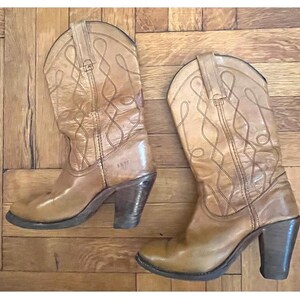 May include: A pair of tan leather cowboy boots featuring a mid-calf height, stacked block heels, and decorative stitched patterns on the shaft. The boots have pull tabs at the top and the brand name Frye is embossed on the side of the leather.
