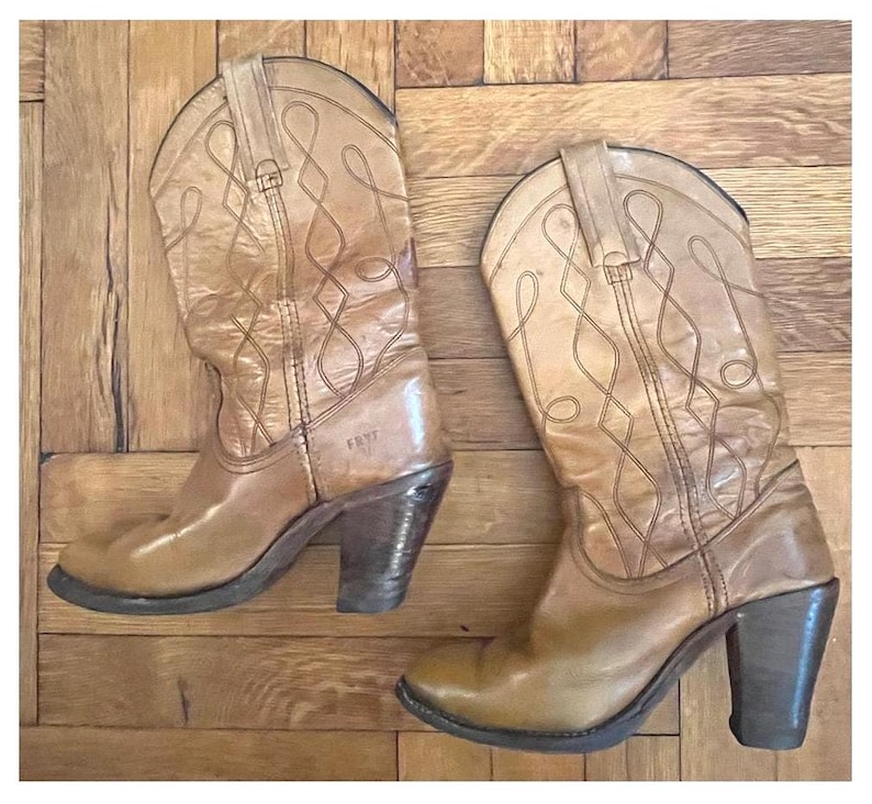 May include: A pair of tan leather cowboy boots featuring a mid-calf height, stacked block heels, and decorative stitched patterns on the shaft. The boots have pull tabs at the top and the brand name Frye is embossed on the side of the leather.
