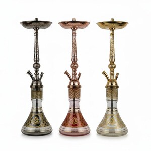 Puede incluir: Tres narguiles decorativos en acabados metálicos: gris oscuro, cobre y dorado. Cada narguile presenta un tallo ornamentado, un cuenco y una base de vidrio con un diseño grabado. Los narguiles se exhiben sobre un fondo blanco.