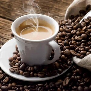 Op de afbeelding: Een witte keramische kop gevuld met dampende hete koffie staat op een bijpassende witte schotel. De kop en schotel zijn omringd door een grote hoop geroosterde bruine koffiebonen, met een metalen schep in een jute zak met bonen.