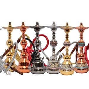 Puede incluir: Una colección de narguiles ornamentados en varios acabados metálicos, incluyendo plata, oro y cobre. Cada narguile presenta una base, un tallo y una manguera decorativos, algunos con acentos coloridos. Los narguiles están dispuestos en fila sobre un fondo blanco.