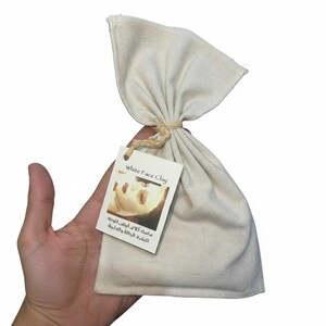 Puede incluir: Una bolsa de lona de color crema atada con hilo, que contiene arcilla blanca para la cara. Una etiqueta adjunta a la bolsa dice "White Face Clay" en inglés y árabe, con una imagen de una persona con una mascarilla facial.