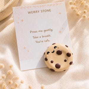 Puede incluir: Una piedra de preocupación con forma de galleta beige con detalles de chispas de chocolate y una cara sonriente, junto a una tarjeta que dice "Worry Stone" y "Press me gently. Take a breath. You're safe."