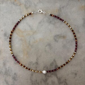May include: Anklet featuring a delicate design with small, colorful beads in shades of gold, burgundy, and silver. A single, round, white pearl is centered on the anklet. The anklet is secured with a silver clasp.