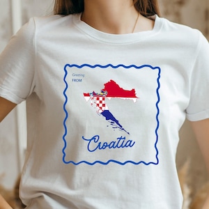 May include: White t-shirt featuring a blue framed graphic of the Croatian map in red, white, and blue. The words "Greeting FROM" and "Croatia" are also in blue.