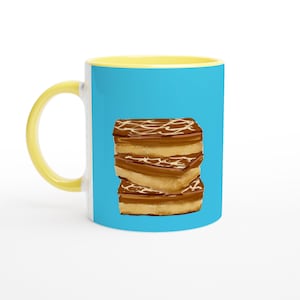 Puede incluir: Una taza de café de cerámica con asa e interior amarillos y exterior azul claro. La taza presenta una pila de tres pasteles rectangulares, marrones y tostados, con glaseado blanco. La taza está sobre un fondo blanco.