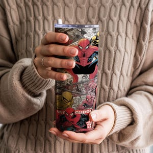 K&ouml;nnte beinhalten: Ein hoher, isolierter Becher mit einer Collage aus Spider-Man-Comics. Das Design zeigt Spider-Man in verschiedenen Posen, mit einem rot-schwarzen Farbschema. Der Becher hat einen silbernen Deckel und einen wei&szlig;en Strohhalm.