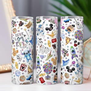 May include: Three white insulated tumblers with colorful Disney-themed illustrations. Each tumbler features various characters, icons, and phrases like "Best Day Ever" and "Never Grow Up". The designs include Stitch, Mickey Mouse, and a Dalmatian.