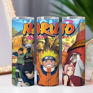 May include: A cylindrical tumbler featuring a colorful graphic wrap of the Naruto anime series. The design displays characters including Naruto, Sakura, and Kakashi against a backdrop of the Hidden Leaf Village and the Hokage mountain monument.