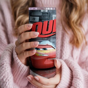 Pu&ograve; includere: Un bicchiere cilindrico in acciaio inossidabile con una stampa grafica di un'auto da corsa rossa con occhi blu e decalcomanie a forma di fulmine giallo. Il design include il testo rosso in grassetto "QU" su sfondo grigio con dettagli.