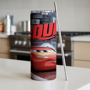 Pu&ograve; includere: Un bicchiere cilindrico in metallo con la grafica di un'auto da corsa rossa con occhi blu e decalcomanie a forma di fulmine giallo. Il design include il testo rosso parziale "QU" su sfondo grigio ed &egrave; dotato di una cannuccia in metallo sottile.