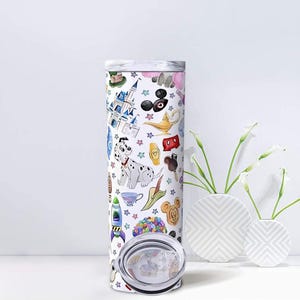 May include: A white, insulated tumbler with a colorful cartoon design featuring a Dalmatian, castle, and other whimsical illustrations. The tumbler has a silver lid and is displayed with white vases and flowers.