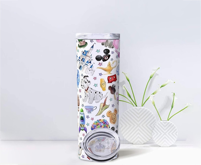 May include: A white, insulated tumbler with a colorful cartoon design featuring a Dalmatian, castle, and other whimsical illustrations. The tumbler has a silver lid and is displayed with white vases and flowers.