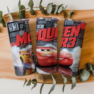 Pu&ograve; includere: Tre bicchieri cilindrici con la grafica di un'auto da corsa rossa con il numero 95, il testo McQueen e uno sfondo di luci da stadio. Il design presenta una finitura metallica, un coperchio trasparente e una tavolozza di colori rosso, nero e grigio.
