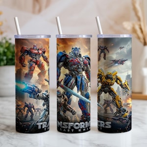 May include: Three stainless steel tumblers with lids and straws, each featuring a different Transformers character. The tumblers have a cityscape background and the text "TRANSFORMERS THE LAST KNIGHT".
