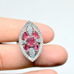 Peut inclure: Bague argentée ornée d'un motif ovale. Elle présente des pierres roses ovales au centre, entourées de petits diamants ronds et transparents. Le design est symétrique et attire le regard. La bague est tenue par une main.