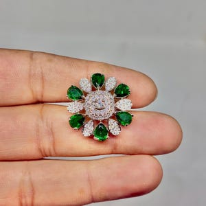 Peut inclure: Broche florale avec un groupe central de pierres claires, entouré d'alternances de pierres vertes en forme de goutte et de petites pierres blanches. La broche est tenue dans une main.