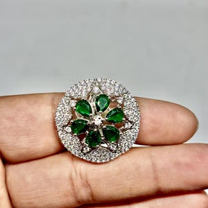 Peut inclure: Une bague argentée ornée d'un motif floral. Le centre de la bague présente un groupe de pierres précieuses vertes vives en forme de goutte. L'anneau extérieur est incrusté de petites pierres précieuses claires, créant un effet de halo étincelant. La bague est tenue par une main.