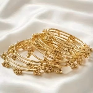 May include: A collection of gold bangles with small, dangling charms, displayed on a white, silky fabric. The bangles feature a delicate design with multiple thin bands and small, round embellishments. The overall aesthetic is elegant and ornate.