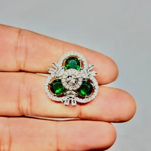 Peut inclure: Une bague argentée avec un motif floral, ornée de trois grandes pierres précieuses rondes vert émeraude. La bague est ornée de pierres plus petites, claires, rectangulaires et rondes, créant un effet scintillant. La bague est tenue dans une main.