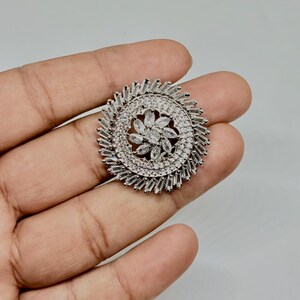 Peut inclure: Une bague argentée avec un motif floral. La bague présente une forme de fleur centrale entourée d'un cercle de petites pierres transparentes. Le bord extérieur présente un motif en forme d'éclat de soleil de pierres rectangulaires.