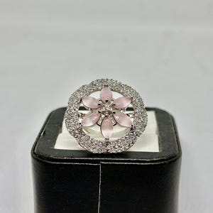 Peut inclure: Bague argentée ornée d'une fleur centrale aux pétales rose clair. La fleur est entourée d'un halo de petites pierres transparentes. La bague est présentée sur un écrin noir.