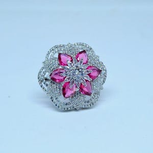 Peut inclure: Bague argentée ornée d'un motif floral. Le centre de la bague est une fleur composée de pierres roses en forme de goutte, entourée de petites pierres transparentes. La monture est également incrustée de pierres transparentes.
