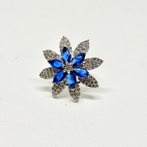 Peut inclure: Bague en forme de fleur avec un anneau argenté. La bague présente un groupe central de pierres bleues entouré d'un cercle de pierres claires et scintillantes. Les pétales sont détaillés, sur fond blanc.