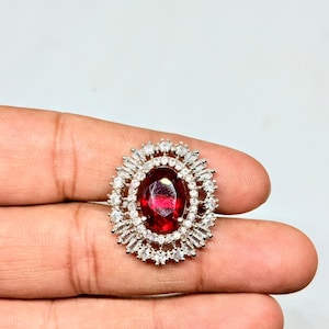 Peut inclure: Une bague en argent ornée d'une grande pierre précieuse ovale rouge foncé au centre, entourée d'un halo de petites pierres claires, rectangulaires et rondes. Le design de la bague est complexe et attrayant, pour les occasions spéciales.