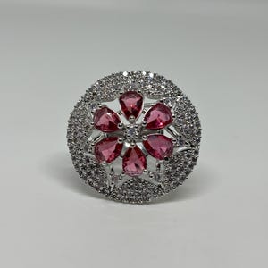 Peut inclure: Bague argentée ornée d'un motif floral. Le centre de la bague présente six pierres roses en forme de goutte, disposées autour d'une pierre centrale. L'anneau extérieur est incrusté de petits cristaux transparents, créant un effet scintillant.