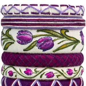 Puede incluir: Una pila de pulseras decorativas en tonos morados, blancos y verdes. Las pulseras presentan patrones florales y geométricos bordados. Los diseños incluyen tulipanes morados y detalles cosidos, creando una apariencia en capas y texturizada.