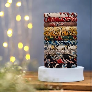 Puede incluir: Una pila de brazaletes cubiertos de tela en varios colores y patrones. Los brazaletes están apilados sobre un pedestal blanco y redondo. Los patrones incluyen diseños florales y abstractos en rojo, marrón, amarillo, azul y negro.