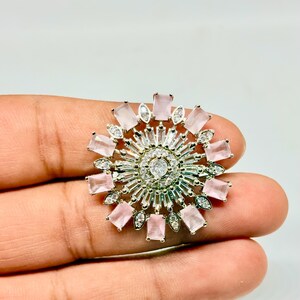 Peut inclure: Broche argentée en forme de fleur, ornée d'une pierre centrale transparente et de pierres roses rectangulaires et transparentes. La broche est tenue à la main sur fond blanc, mettant en valeur son design complexe et ses détails scintillants.