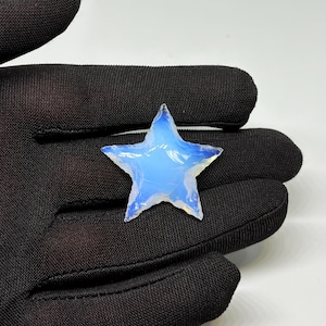 May include: A star-shaped gemstone with a milky, opalescent appearance and a pale blue colour. The stone is held in a black glove against a white backdrop. The edges of the star are slightly uneven, giving it a natural look.